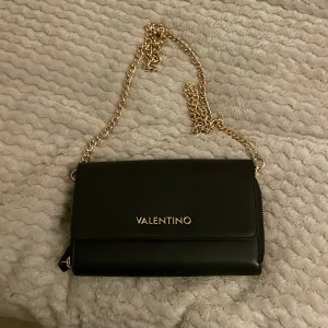 Svart axelväska från Valentino - Snygg svart axelväska från Valentino med guldkedja som axelrem. Väskan har en stilren design med logotyp framtill och dragkedja inuti för säker förvaring. Perfekt för att bära dina viktigaste saker med stil. 