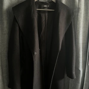 Svart kappa från BikBok - Snygg svart brun grå kappa från BikBok i ullblandning. Den har en elegant design med knappar framtill och ett bälte i midjan för en stilren look. Perfekt för kyligare dagar.