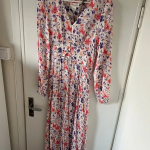 Blommig långklänning från Lindex - Säljer en vacker långklänning från Lindex med blommigt mönster i rött, blått och lila. Färgerna är ljusare än bilden visar. Klänningen har långa ärmar och en v-ringad hals. Aldrig använd, prislappen kvar. Köpt ny för 600kr. 