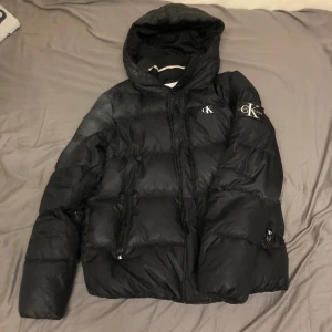 Svart dunjacka från Calvin Klein Jeans - Säljer en svart dunjacka från Calvin Klein Jeans med huva och dragkedja. Jackan har en stilren design med CK-logga på bröstet och ärmen. Perfekt för kyliga dagar.