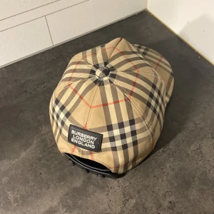 Rutig keps från Burberry - Snygg rutig keps från Burberry i klassiskt beige, svart och rött mönster. Kepsen har en justerbar rem baktill och en broderad Burberry-logga. Perfekt för en stilren look.