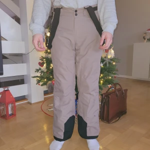 Beige täckbyxor med svarta detaljer - Säljer ett par beige skidbyxor med svarta förstärkningar vid bensluten. Byxorna har justerbara hängslen och dragkedjor vid fickorna. Perfekta för vinterns kalla dagar. Använda en vecka i skidbacken. Finns ett mindre svart märke på ena benet. Går antagligen bort i tvätt.