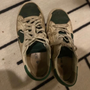 Vita och gröna sneakers från The Gallerist - Snygga vita sneakers med gröna detaljer från The Gallerist. Skorna har en unik design med gröna klöver på sidan och text på hälen. Perfekta för en avslappnad stil. 