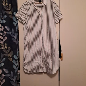 Vit och svart randig skjortklänning - Säljer en stilren vit skjortklänning med svarta ränder. Klänningen har korta ärmar och en klassisk krage. Perfekt för en avslappnad och chic look. Passar bra till både sneakers och sandaler.