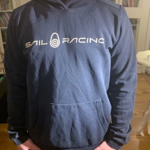 Mörkblå hoodie från Sail Racing - Säljer en mörkblå hoodie från Sail Racing med vit logga på bröstet. Tröjan har en klassisk känguruficka och långa ärmar. Perfekt för en avslappnad stil.