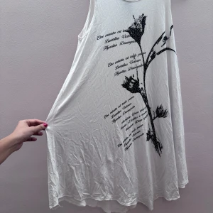 Vit ärmlös klänning med tryck - Säljer en vit ärmlös klänning med ett svart blommönster och texttryck på framsidan. Klänningen har en luftig passform och är perfekt för varma dagar. Den är gjord i ett mjukt och bekvämt material.