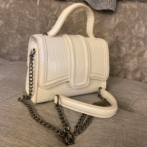 Vit handväska från Zara - Säljer en superfin vit handväska från Zara. Den har en snygg krokodilpräglad yta och en kedja som axelrem. Perfekt för att ge din outfit en chic touch! Väskan är i gott skick förutom någon form utav missfärgning på vissa kanter, går kanske att tvätta bort men är ingenting som syns vid användning. Praktisk storlek för alla dina nödvändigheter.