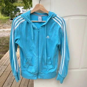 Blå hoodie från Adidas - Säljer en snygg och bekväm ljusblå hoodie från Adidas. Den har de klassiska tre vita ränderna längs ärmarna och en dragkedja framtill.  Hoodien är i bra skick men förekommer dock några fläckar och ett litet hål se bild. 