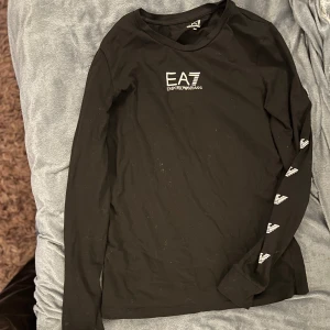 Svart långärmad tröja från Emporio Armani - Säljer en stilren svart långärmad tröja från Emporio Armani. Tröjan har deras ikoniska logga tryckt på bröstet och längs ärmarna. Perfekt för en casual look eller att klä upp med accessoarer. Passar både till vardags och fest!