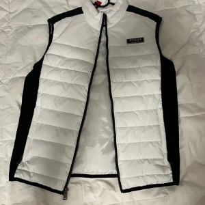 Vit och svart dunväst från Guess - Säljer en snygg och stilren vit och svart dunväst från Guess. Västen har en dragkedja framtill och är perfekt för kyligare dagar. Den är i storlek XS och har en klassisk design med Guess-loggan på bröstet. Perfekt för både vardag och lite finare tillfällen! köpt för 1200 från guess och är i nyskick och aldrig anväd.