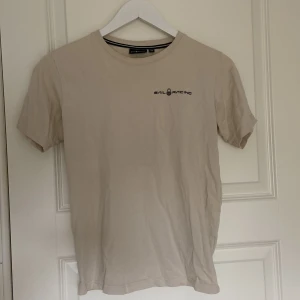 Beige t-shirt från Sail Racing - Säljer en snygg beige t-shirt från Sail Racing. Den är i storlek 160 och har en enkel design med loggan tryckt på bröstet.T-shirten är kortärmad och i bomull, vilket gör den skön att ha på sig. Passar bra till både jeans och shorts!