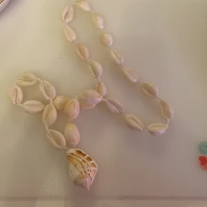 Halsband med snäckor - Säljer ett supercoolt halsband med vita snäckor. Det är perfekt för sommaren och ger en härlig strandkänsla. Snäckorna är jämnt fördelade och det finns en större snäcka som hänger som en berlock. Passar perfekt till en bohemisk stil eller för att ge en outfit en naturlig touch.