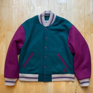 Grön och lila bomberjacka från Acne Studios - Snygg bomberjacka från Acne Studios i en cool kombination av grönt och lila. Jackan har långa ärmar och stängs med knappar. Den har randiga muddar i beige, lila och rött samt quiltat foder för extra värme. Perfekt för att sticka ut i mängden!