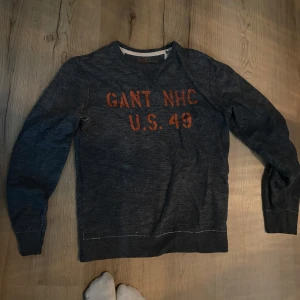 Ljusgrå tröja från GANT - Säljer en ljusgrå tröja från GANT med texten 'GANT NHC U.S. 49' i orange på framsidan. Tröjan har långa ärmar och en rund halsringning. Perfekt för en avslappnad stil.
