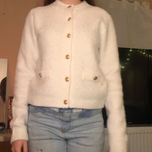 Vit kofta från Holly & Whyte - Säljer en stilren vit kofta från Holly & Whyte by Lindex. Koftan har dekorativa guldknappar och två fickor framtill. Perfekt för en elegant och mysig look.