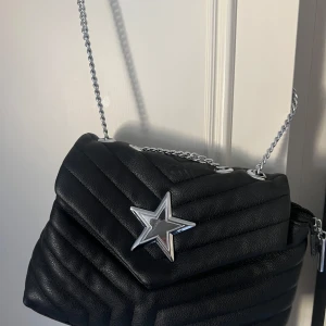 Svart quiltad axelväska med stjärndetalj - Snygg svart axelväska i quiltat mönster med en cool silverfärgad stjärndetalj på framsidan. Väskan har en kedjerem och flera fack inuti för smart förvaring. Perfekt för att ge din outfit en edgy touch!