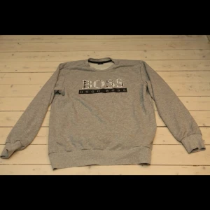 Grå sweatshirt från Hugo Boss - Säljer en stilren grå sweatshirt från Hugo Boss med deras ikoniska logga tryckt på bröstet. Tröjan har långa ärmar och ribbade muddar vid ärmslut och nederkant. Perfekt för en avslappnad look.