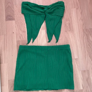 Grön kjol med matchande tubtopp - Snygg grön kjol med matchande tubtopp. Kjolen har en kort längd och ett subtilt ribbat mönster som ger en cool textur. Toppen är en tubtopp med knytning framtill, vilket ger en trendig look. Perfekt för sommarens äventyr!