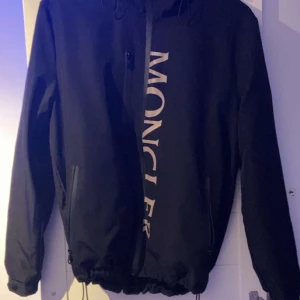 Svart vindjacka från Moncler - Snygg svart vindjacka från Moncler med dragkedja och huva. Jackan har långa ärmar och Moncler-logga på bröstet samt en liten logga på ärmen. Perfekt för blåsiga dagar.