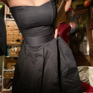 Svart klänning från Vila - svart klänning från Vila i storlek XS. axelbandslös men finns öglor om man vill lägga till egna. volangdetalj i tyll vid fållen. ganska liten i bysten. går ungefär till lite över knäna. stängs med dragkedja i sidan. 