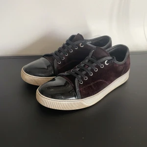 Vinröda skor från Lanvin - Snygga svarta sneakers från Lanvin med en stilren design. Skorna har en kombination av mocka och glansigt material på tån. De har snörning och en vit sula som ger en modern touch. Perfekta för en trendig look. Skick 7,5! Vid frågor är det bara att höra av sig! Liten skada på Captoen.