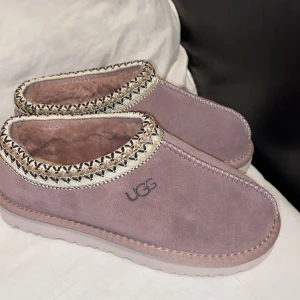 Rosa tofflor från UGG - Mysiga rosa tofflor från UGG med mjukt foder och dekorativ kant. Perfekta för att hålla fötterna varma och bekväma. Snygg design med UGG-logga på sidan och mönstrad sula för bra grepp.