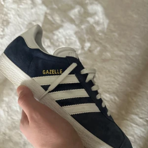 Adidas Gazelle sneakers i blått och vitt - HELT NYA.Snygga Adidas Gazelle sneakers i blå mocka med vita detaljer och snörning. Den klassiska designen har tre vita ränder på sidan och en vit sula. Perfekta för en stilren look. Använa ett fåtal gånger.
