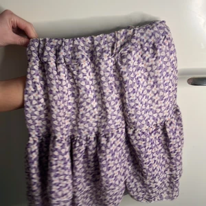 Lila blommig kjol från Homlys - Charmig lila kjol från Homlys med ett blommigt mönster i vitt och rosa. Kjolen har en volangdetalj som ger den en lekfull touch. Perfekt för en somrig look!