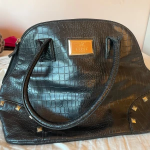 Svart handväska med krokodilmönster - Snygg svart handväska med krokodilmönster och gulddetaljer . Väskan har två handtag och dekorativa nitar i hörnen. Perfekt för att ge din outfit en elegant touch.
