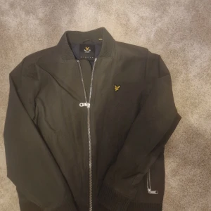 Militär grön bomberjacka från Lyle & Scott - Säljer en stilren svart bomberjacka från Lyle & Scott med dragkedja och ribbade detaljer. Jackan har en broderad logotyp på bröstet och är långärmad. Perfekt för en cool och avslappnad look.