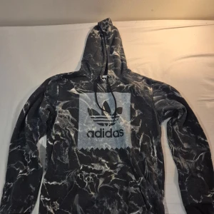 Svart och vit mönstrad hoodie från Adidas - Säljer en grå och vit mönstrad hoodie från Adidas med det klassiska logotyptrycket på framsidan. Hoodien har en justerbar huva och långa ärmar, perfekt för en avslappnad stil.