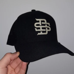 Svart keps med broderad logga - Snygg svart keps från Dijnns med en broderad logga framtill. Kepsen har en klassisk design med nätpaneler baktill för extra ventilation. Perfekt för en cool och avslappnad stil. Oanvänd utan kvitto! 