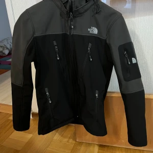 Svart jacka från The North Face - Säljer en svart jacka från The North Face i storlek S. Jackan har flera praktiska dragkedjefickor och en justerbar huva. Perfekt för utomhusaktiviteter med sin stilrena design och funktionalitet.