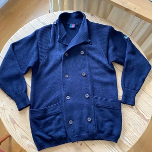 Mörkblå dubbelknäppt cardigan med knappar 100%ull - Säljer en stilren mörkblå kofta/cardigan med dubbelknäppning och ribbade detaljer på axlarna. Koftan har långa ärmar och praktiska fickor framtill. Perfekt för kyligare dagar! 100% ull o vintage från 70tal/80tal o sååå bra kvalitet!! Skulle säga herr storlek M/L