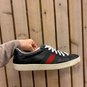 Svarta guuci skor - Snygga svarta sneakers med vita snören och en klassisk röd och grön rand på sidan. Skorna har en vit sula och är tillverkade i skinn med ett diskret mönster. Perfekta för en stilren look.