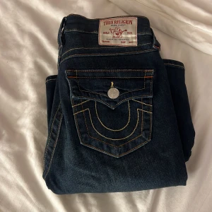 Mörkblå True Religion jeans - Säljer ett par mörkblå jeans från True Religion med ikoniska kontrastsömmar och snygga detaljer på bakfickan. Klassisk femficksmodell med knappgylf och silverfärgade knappar. Jeansen har en rak passform och är tillverkade i slitstarkt denimtyg.