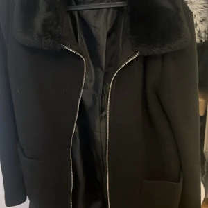 Svart kappa med fuskpäls - Snygg svart kappa med mjuk fuskpäls på kragen och dragkedja framtill. Perfekt för kyliga dagar när du vill hålla stilen. Den har en klassisk look som funkar till många tillfällen.