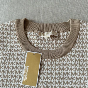 Michael Kors set kjol+tröja - Säljer ett snyggt beige set från Michael Kors med ett stilrent mönster i brunt. Tröjan har korta ärmar och ribbade kanter vid hals, ärmslut och nederkant. Kjolen är till knäna och thight då den formar kroppen. Super bra material, drar in kroppen och formar den. Aldrig använd. Tröja i M och kjolen i S. Säljs tillsammans Perfekt för en trendig look!