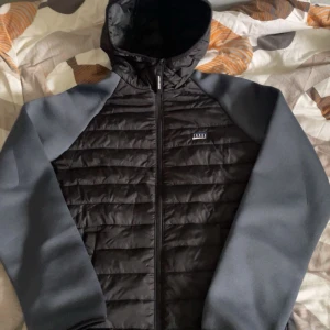  jacka från Jack & Jones - Säljer en snygg svart och grå jacka från Jack & Jones. Jackan har en quiltad design med dragkedja och huva. Perfekt för kyligare dagar med sin stilrena look och bekväma passform.