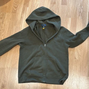 Grön hoodie från Ralph Lauren - Säljer en grön hoodie från Ralph Lauren med dragkedja och broderad logga på bröstet. Tröjan har långa ärmar och en praktisk huva. Perfekt för en avslappnad stil.
