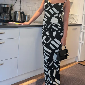 Svartvit mönstrad långklänning - Snygg svartvit långklänning med geometriskt mönster. Klänningen har smala axelband och en figurnära passform som framhäver silhuetten. Perfekt för en stilren och modern look.