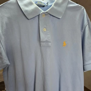 Ljusblå pikétröja från Ralph Lauren - Snygg ljusblå pikétröja från Ralph Lauren med klassisk krage och knappar. Den har en broderad guld logotyp på bröstet. Perfekt för en stilren look.