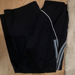 Mörkblåa träningsbyxor med mönster - Snygga mörkblåa träningsbyxor med vitblåa ränder längs sidorna och elastisk midja. Liknar parachute pants. Står inget märke eller storlek men skulle säga dem är storlek S/M. Köpt på plick men har aldrig använt. 