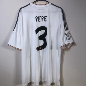 Vit Real Madrid fotbollströja - Vit Real Madrid fotbollströja med korta ärmar och spelarnamnet 'Pepe' samt nummer 3 på ryggen. Tröjan har svarta detaljer och LFP-logga på ärmen.