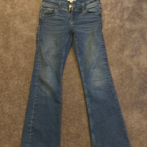 Blå bootcut jeans - Snygga blå bootcut jeans med hög midja och klassisk femficksdesign. Jeansen har en dragkedja och knappar framtill samt dekorativa knappar på bakfickorna. Perfekta för en avslappnad stil.