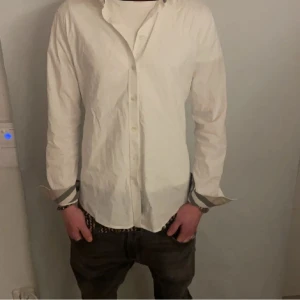 Burberry - Snygg vit skjorta med långa ärmar och stilrena knappar. Skjortan har unika randiga detaljer på insidan av ärmsluten som ger en extra touch. Perfekt för en stilren look. Knappt använd skick 9/10