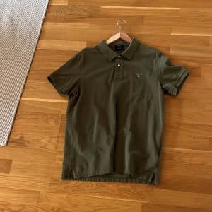 Olivgrön pikétröja från Gant - Snygg olivgrön pikétröja från Gant med klassisk krage och knappar framtill. Perfekt för en avslappnad men stilren look. Tröjan har korta ärmar och en liten broderad logga på bröstet.