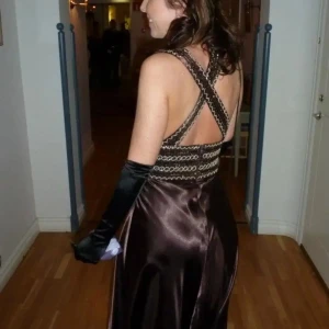 Balklänning  - Elegant brun långklänning med v-ringad hals och dekorativa band i midjan. Klänningen har en öppen rygg och är tillverkad i ett glansigt material som ger en lyxig känsla. Perfekt för speciella tillfällen.