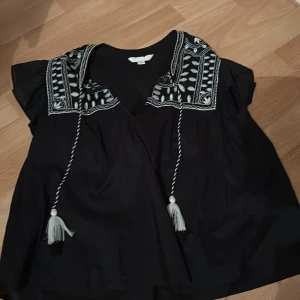 Svart blus med broderi och knytband - Charmig svart blus med vackert vitt broderi på axlarna och knytband med tofsar framtill. Blusen har korta ärmar och en avslappnad passform, perfekt för en bohemisk look.