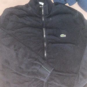 Svart tröja med dragkedja från Lacoste - Säljer en svart tröja från Lacoste med dragkedja och det klassiska krokodilmärket på bröstet. Tröjan har långa ärmar och ribbade muddar. Perfekt för en stilren look.